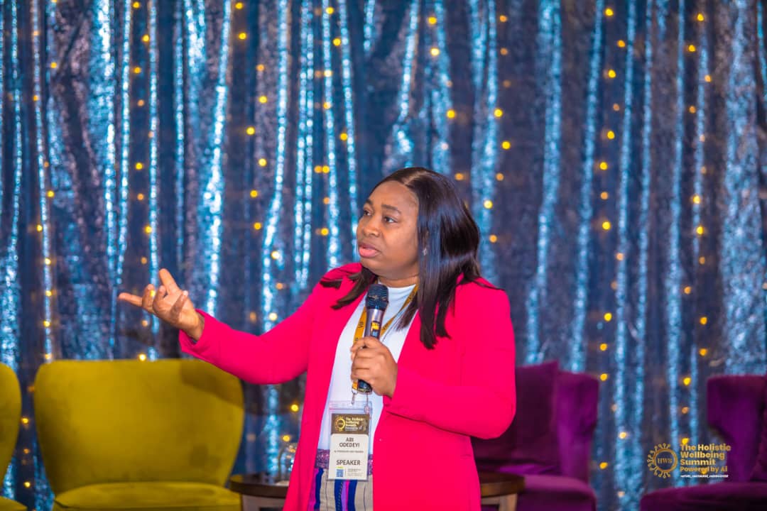 Abi Odedeyi in a red blazer speaking with a microphone on a lit stage at the Holistic Wellbeing Summit powered by AI